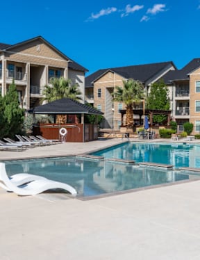 A large swimming pool surrounded by lounge chairs and umbrellas in front of apartment buildings.