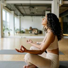 A woman is sitting cross-legged on a yoga mat in a room with a high ceiling and large windows.