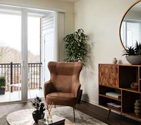 A brown chair is in front of a large window in a room with a wooden shelf and a round mirror.