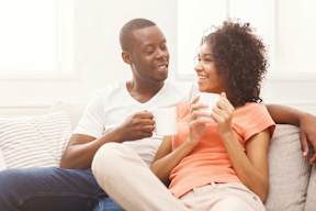 A man and a woman are sitting on a couch, each holding a cup and smiling at each other.