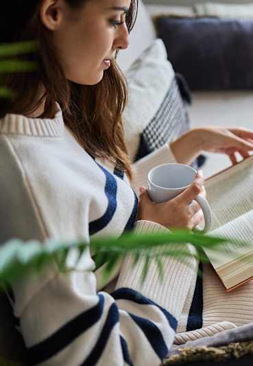 A woman is sitting on a couch reading a book and holding a cup.