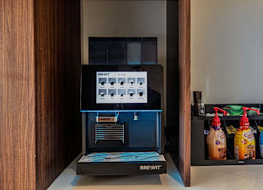 A Brewit coffee machine is on a counter with a shelf of drinks beside it.