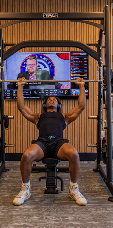 A man is working out in a gym with a television mounted on the wall behind him.