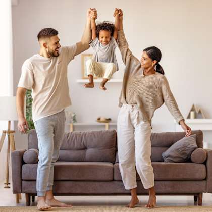 a man and woman standing in a living room with a child jumping on a couch