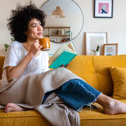 a woman with curly hair is sitting on a yellow couch, holding a mug and reading a book.