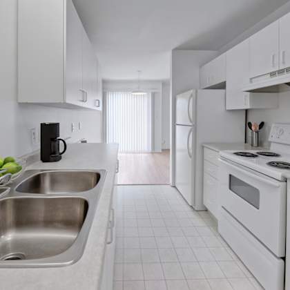 a clean galley-style kitchen here at willowview terrace with white cabinets, a double stainless-steel sink, and bright countertops for a simple modern look. a white refrigerator and electric stove sit along the right side, with light flooring and a dining area visible near the sliding glass doors at the back.