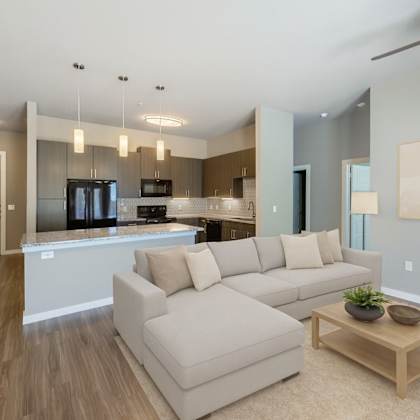 a bright, open-concept living area here at harmony luxury with a modern sectional sofa, light wood coffee table, and soft neutral décor, flowing into a stylish kitchen with dark cabinetry, granite countertops, pendant lighting, and sleek black appliances for a contemporary, upscale feel.