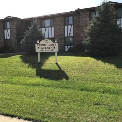 a sign for carrie lawn apartments stands in the grass in front of a brick building.