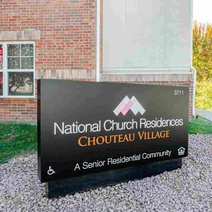 a sign for national church residences chouteau village, a senior residential community, is displayed in front of a brick building.