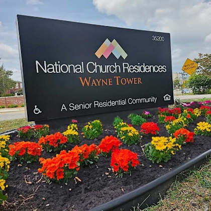 a sign for national church residences wayne tower, a senior residential community, is displayed in front of a flower bed.
