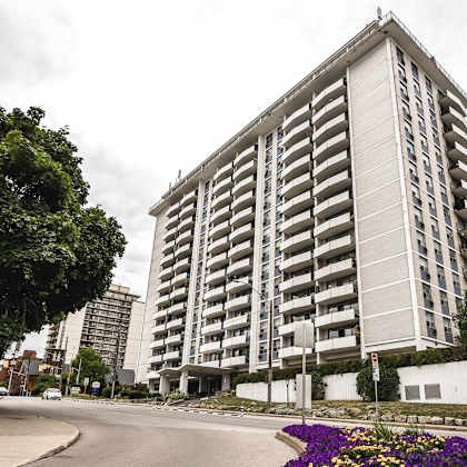 a tall residential building with balconies is surrounded by trees and a street with a flower bed.