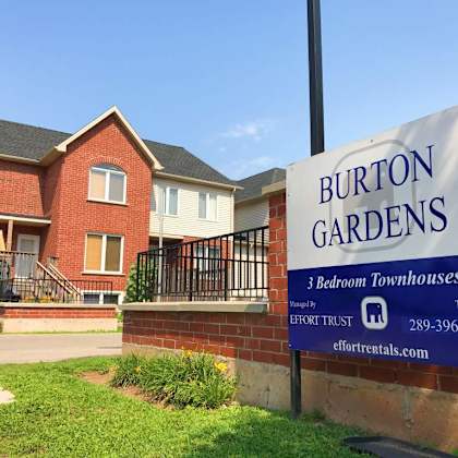 a sign for burton gardens 3 bedroom townhouses stands in front of a brick house.
