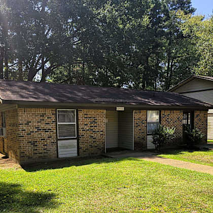 a small brick house with a brown roof and a white door is surrounded by green grass and trees.