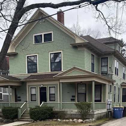 a green two-story house with a porch and a tree in front.