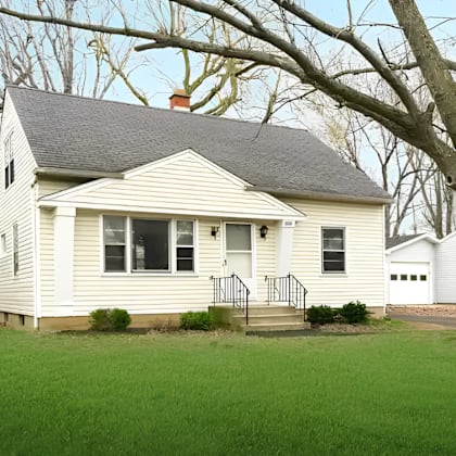a small white house with a grey roof and a tree in front.