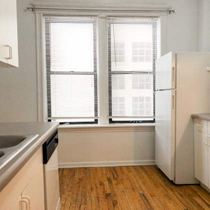 a kitchen with a white refrigerator, wooden floors, and a window with blinds.