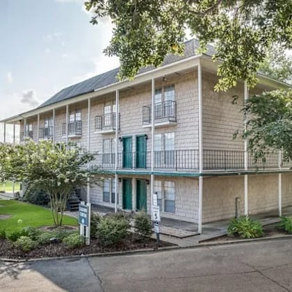 a building with a balcony and a green door is surrounded by trees and has a parking lot in front.