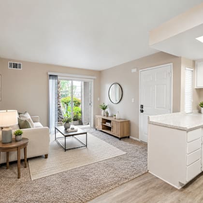 a well-lit, spacious living room with a white sofa, a wooden side table, and a large window.