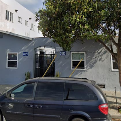 a grey car is parked in front of a grey building with a tree in front of it.