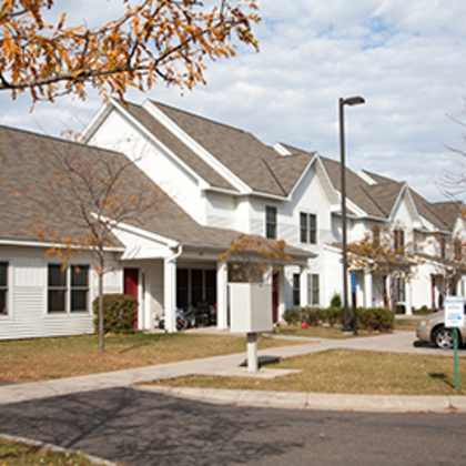 a white house with a grey roof and a tree with orange leaves in front.