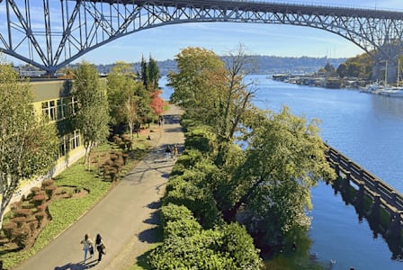 Two people walking on a path under a bridge over a river.