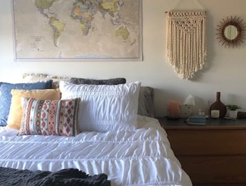White bedspread with a white pillow and a brown and pink patterned pillow.  Large map on wall behind bed with dresser to right of bed.