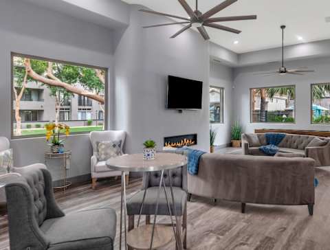 A living room with a grey couch, a white armchair, a grey chair, a round table, a television, a fireplace, and a ceiling fan.