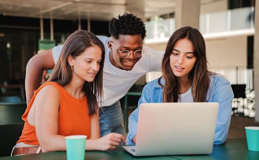 Three people are gathered around a laptop, with one person standing and pointing at the screen.