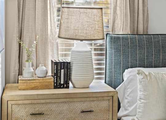 A lamp with a beige shade sits on a wooden nightstand next to a blue striped pillow at West Oak Apartments, North Carolina