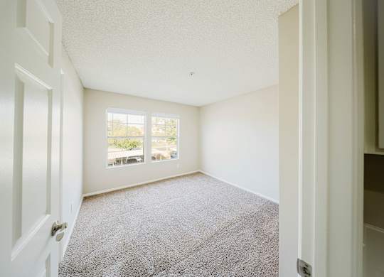 A room with a carpeted floor and a window with a view of trees and a balcony. at Legends at Rancho Belago, Moreno Valley, CA, 92553