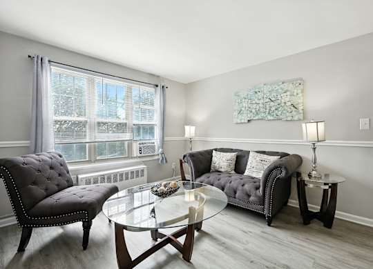 A living room with a grey couch, a black chair, and a glass coffee table at Huntwood Courts Apartments, Washington DC