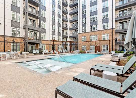 A large outdoor pool area with sun loungers and umbrellas in front of apartment buildings at Hawkins Press Apartments, North Carolina, 28203