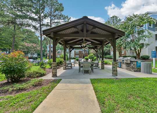 A pavilion with a concrete floor and a roof made of wood and metal is surrounded by trees and bushes.