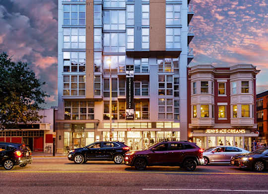 A modern building with a glass facade is illuminated from within, with cars parked in front and a street scene in the foreground.