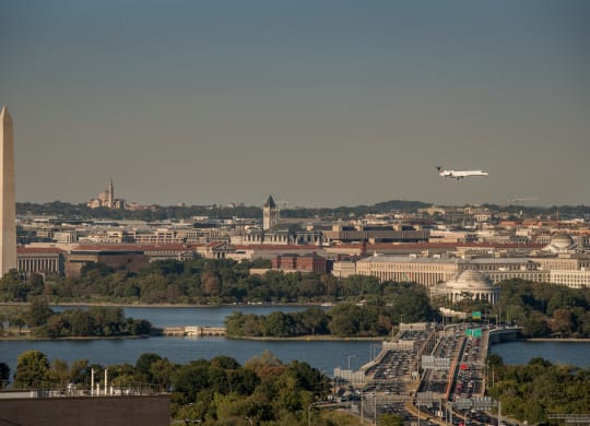a plane flies over the capital of the united states with the washington monument in the foreground at The Acadia at Metropolitan Park, Arlington, 22202