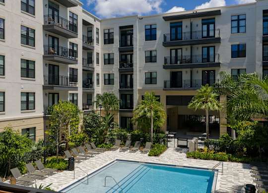 A swimming pool is surrounded by palm trees and lounge chairs in front of apartment buildings.