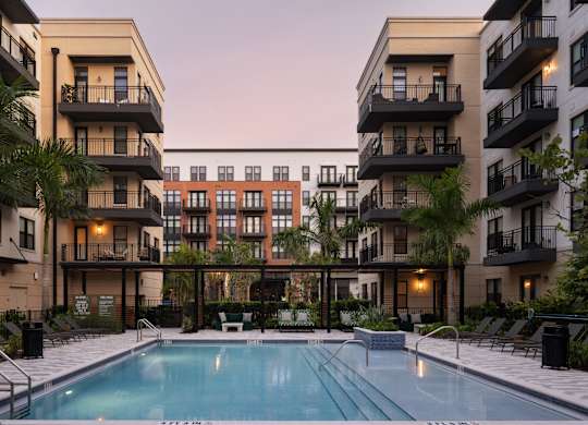 A swimming pool is surrounded by lounge chairs and palm trees in front of apartment buildings.