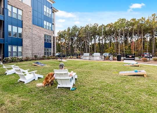 A person is sitting on a white chair in a grassy area with a dog and a picnic table.