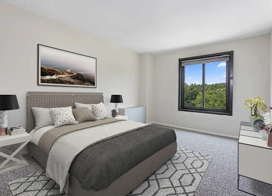 A bedroom with a large bed, a nightstand with a lamp, a framed picture on the wall, and a window with a view of trees.