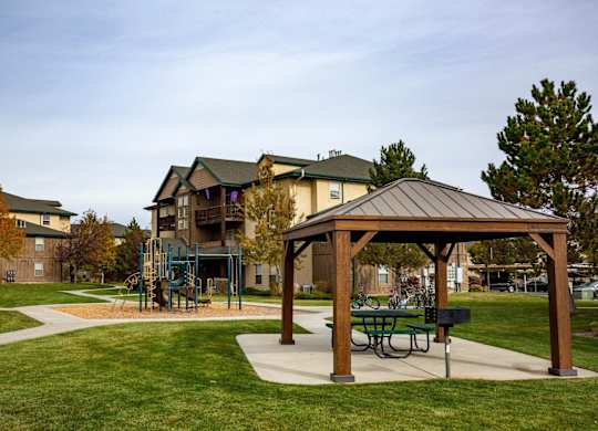 A wooden pavilion with a picnic table sits in a grassy area in front of apartment buildings.at Sunset Ridge, West Jordan