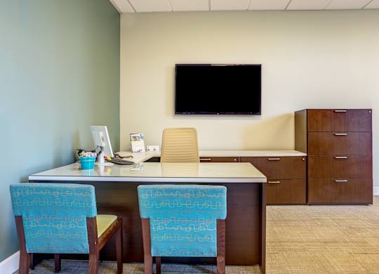 A room with a desk, chairs, and a television mounted on the wall.