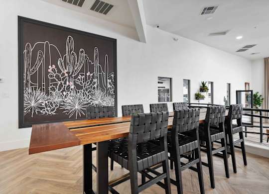 A long wooden table is surrounded by chairs in a room with a desert painting on the wall.