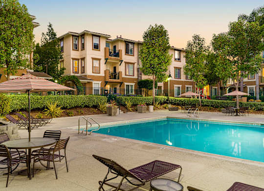 A pool surrounded by chairs and umbrellas in front of apartment buildings.