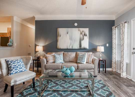 A living room with a grey couch, a blue and white patterned pillow, a glass coffee table, and a painting on the wall.