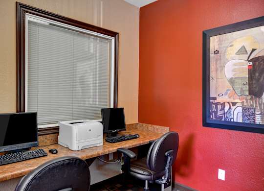A desk with a computer monitor, keyboard, and printer in front of a window with blinds.