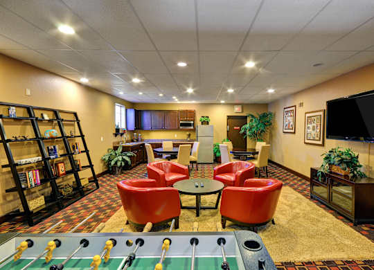 A foosball table is in the middle of a room with red chairs and a bookshelf.