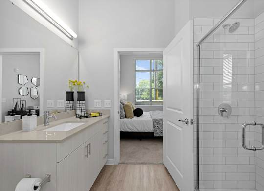 A white bathroom with a walk-in shower and a bed in the background.