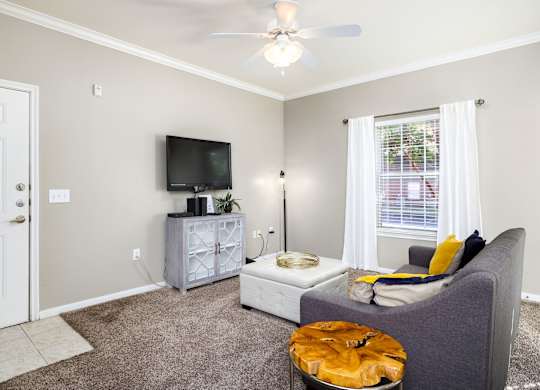 A living room with a grey couch, a television, a fan, and a window with white curtains at Clear Creek Meadows Apartments, Texas