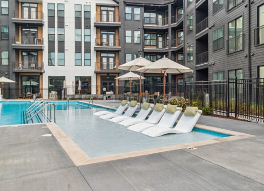 a pool with white lounge chairs in front of an apartment building