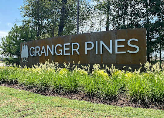 A sign that says "Granger Pines" is surrounded by grass and plants.